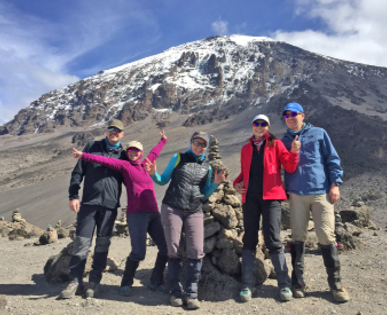 A team of happy climbers pose with the summit of Mount Kilimanjaro clear from cloud behind them.