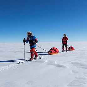 Skiers traverse the great white expanse of interior the Antarctic while hauling sleds, their bright red clothing and orange sleds contrasting against the white now and blue sky.