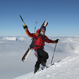 A backcountry ski tourer waves out to the camera as they approach a summit with towering peaks piercing low cloud below.