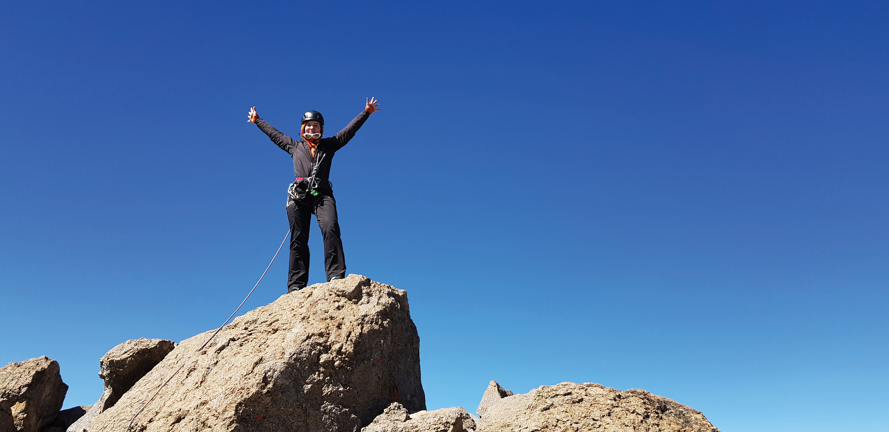 A climber celebrates on the summit of Mount Kenya, cloudless blue skies behind