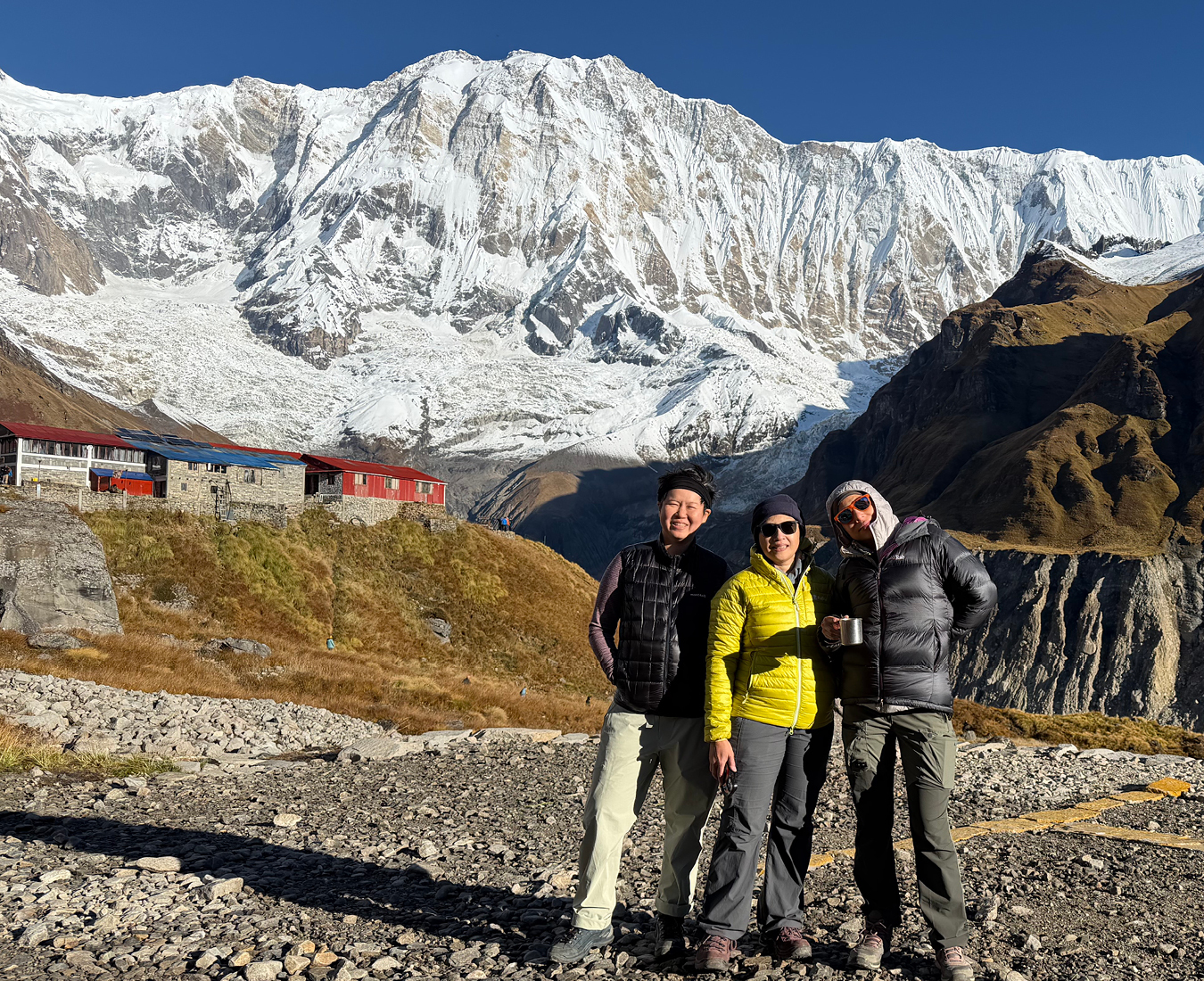 Three trekkers stand on a heli pad at Annapurna Base Camp with 8000m peaks behind and bright blue sky.
