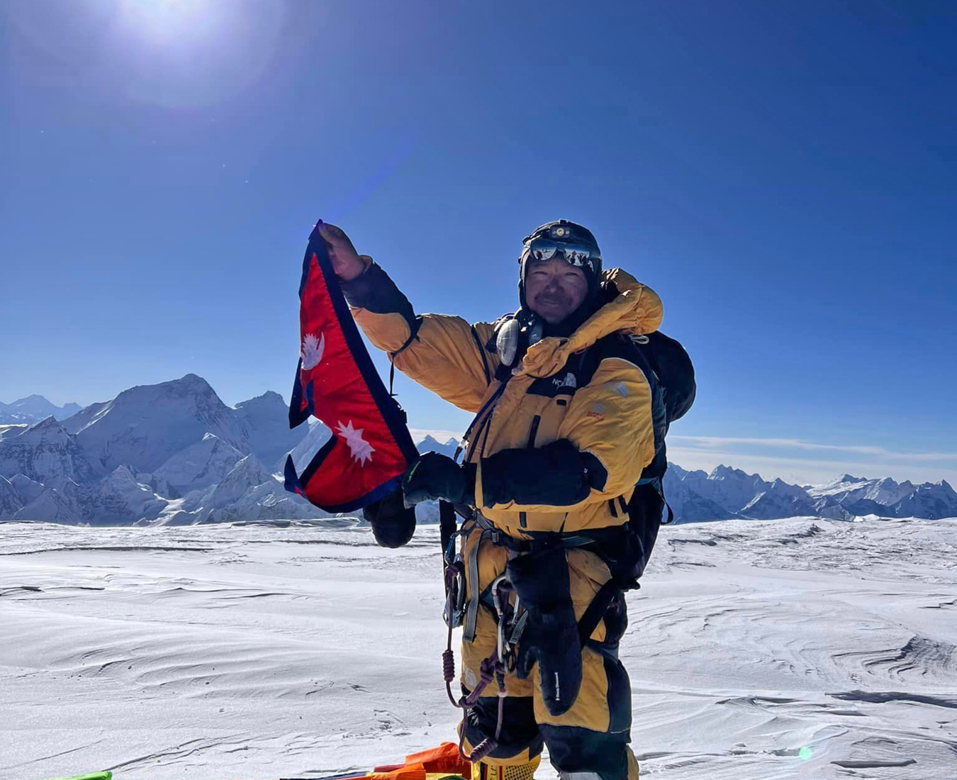 AC Climbing Sherpa Sanduk proudly waves the Nepalese flag on the wide summit plateau of Cho Oyu under bright blue sky with the Himalayan range in the distance.