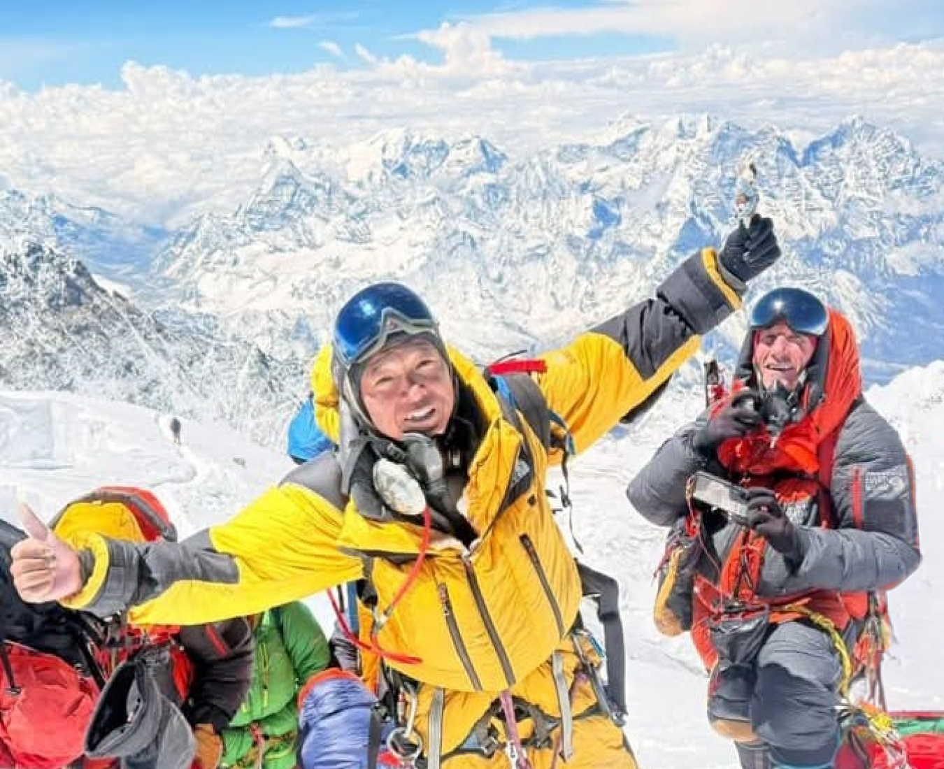 AC Climbing Sherpa Sanduk Tamang in a yellow down suit, arms outstretched with a smiling AC Guide Rob Smith looing on. The vast expanse of the Himalaya behind them into the distance.