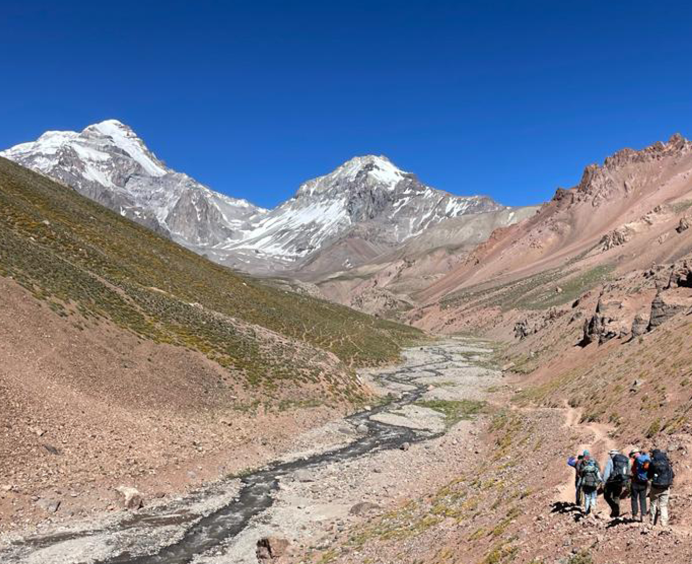 A group treks through the rocky, barren Vacas Valley enroute to Plaza Argentina Base Camp with a snow capped Mount Aconcagua in the distance.