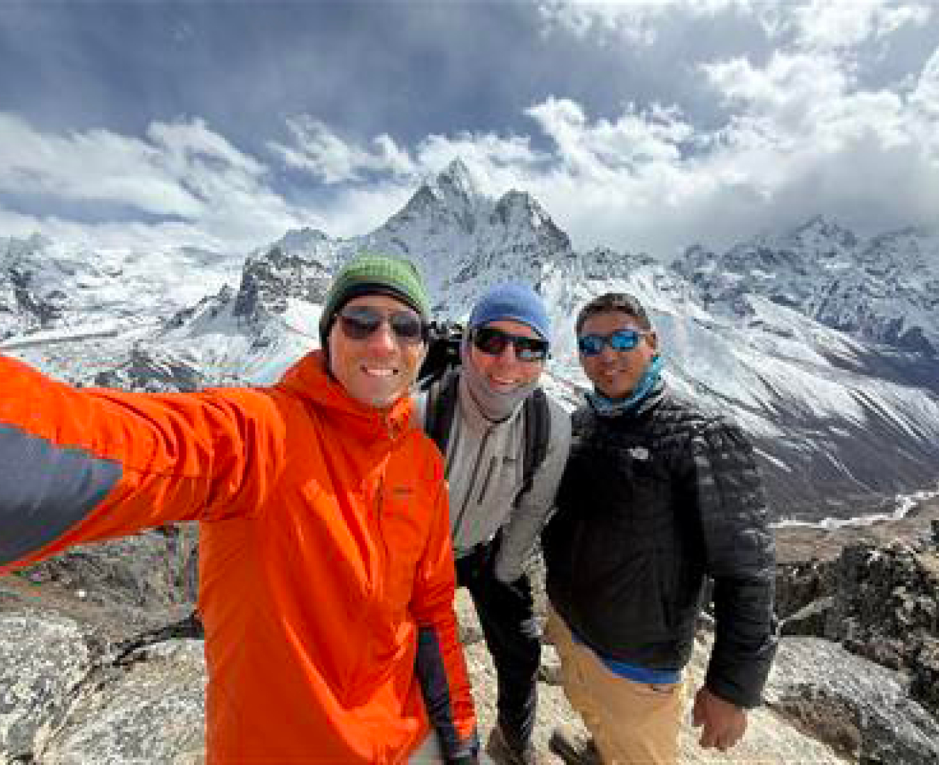Our Island Peak team pose for a selfie on the Kongma La a bright red jacket contrasting with the snow covered Mt Ama Dablam in the background and dramatic clouds above.