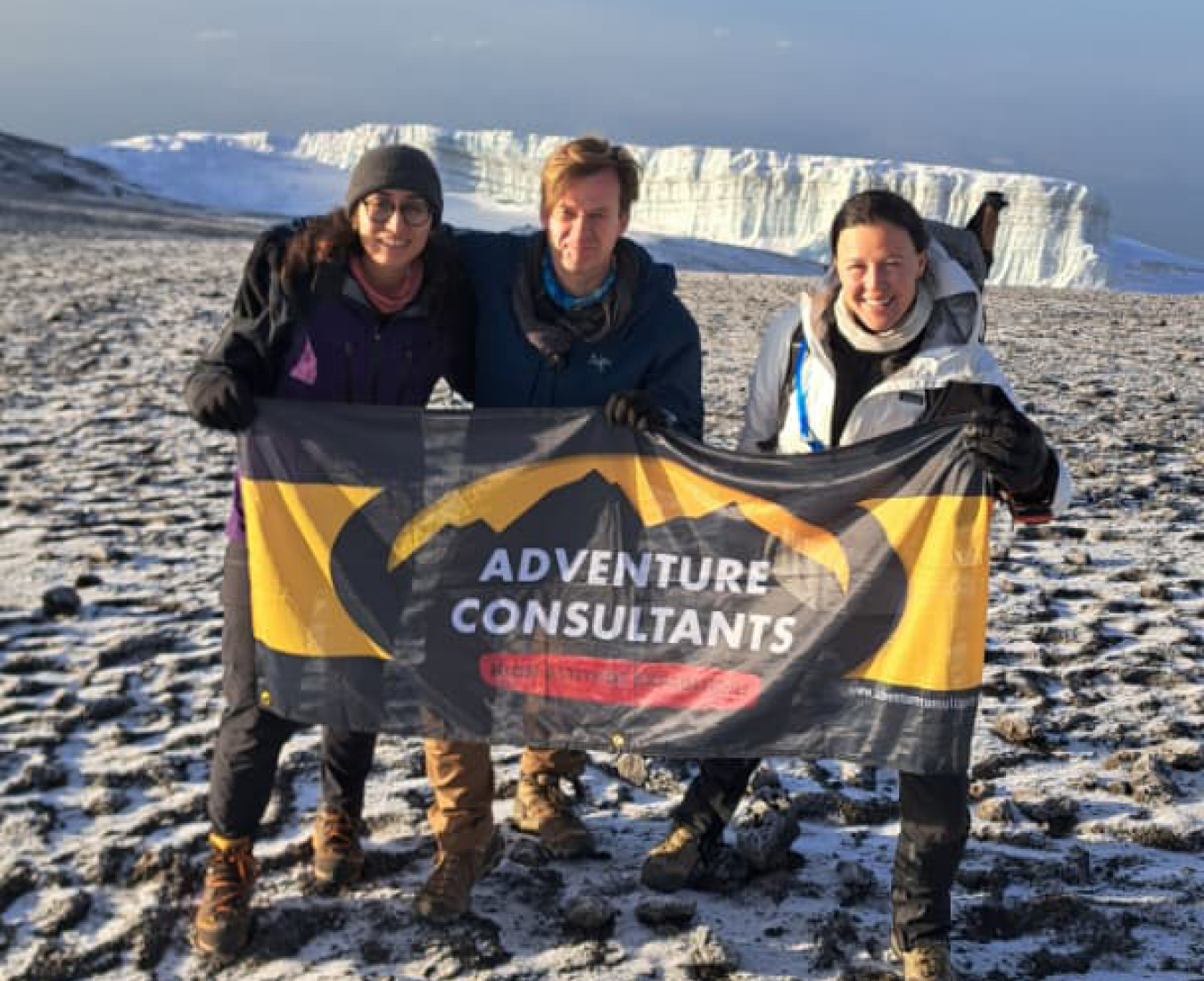AC team on the summit of Mount Kilimanjaro with the summit glacier behind them and the team waving an Adventure Consultants flag.