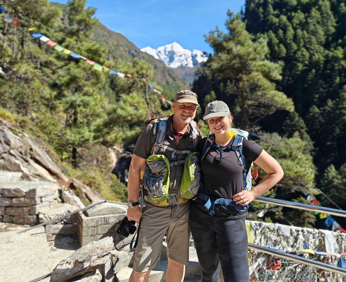 A father and daughter stand alongside the Hillary Bridge near Namche with forest clad hillsides surrounding them and snow covered mountains in the distance.