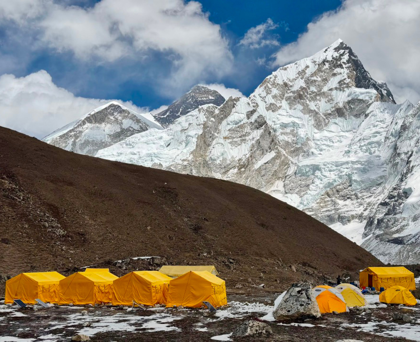 Views of Mount Everest and Nuptse from the private Adventure Consultants camp in Gorak Shep, bright yellow tents in the foreground and blue sky with a few clouds above the mountains.