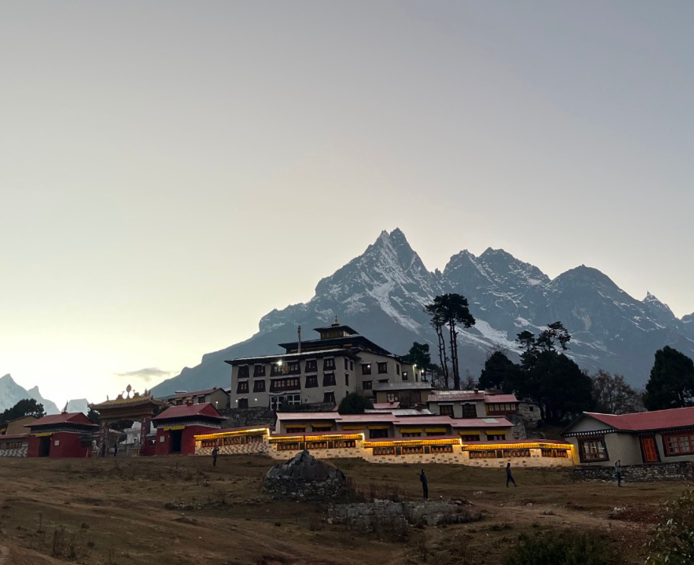 Tengboche Monastery at dusk, its warmly lit buildings glowing against a darkening sky. The jagged, snow-dusted peak of Thamserku rises dramatically behind, with a grassy meadow and scattered trees in the foreground. Khumbu Valley, Nepal.