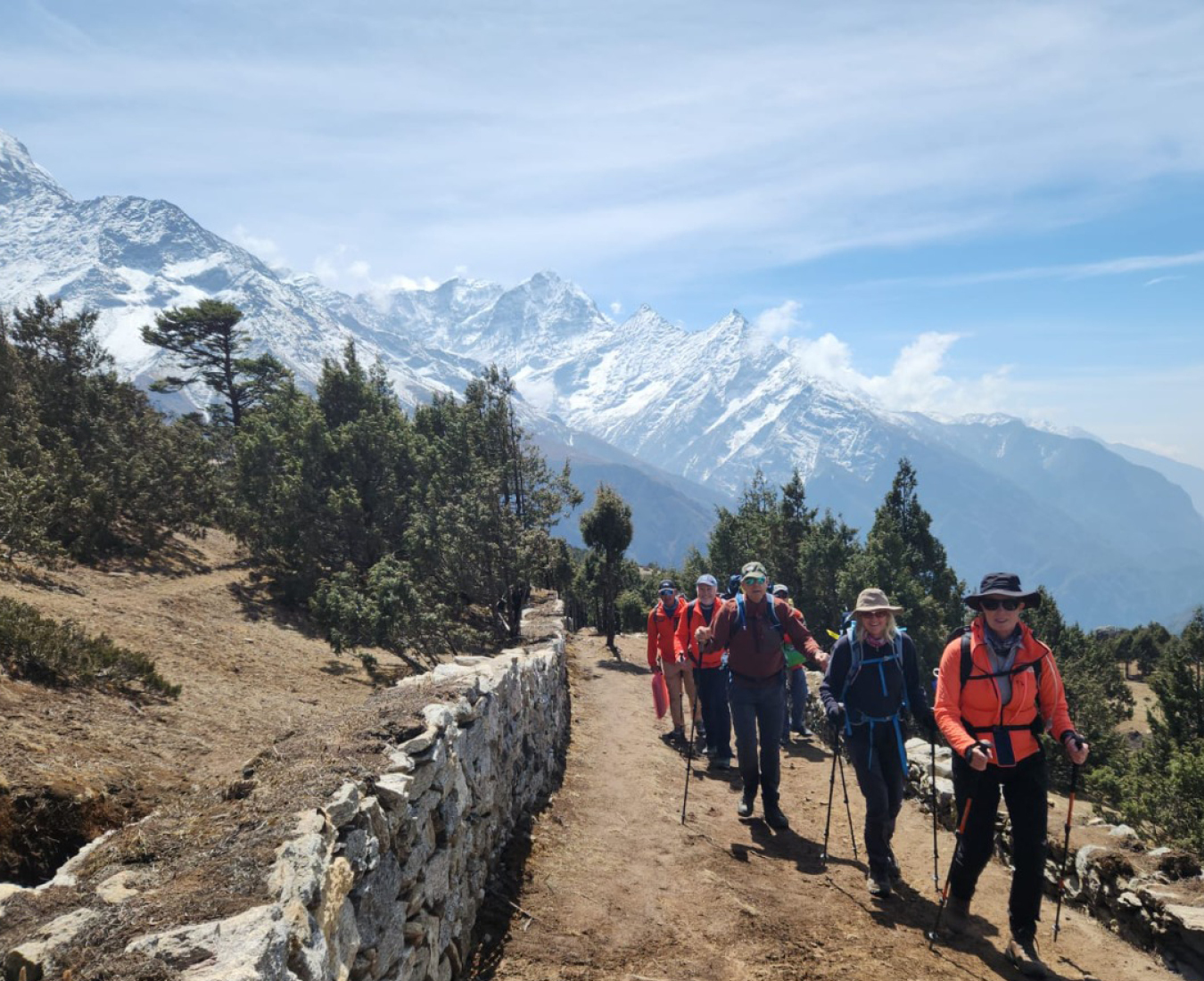 A team treks above Namche village in clear conditions, vegetation around them and snow capped peaks in the distance.