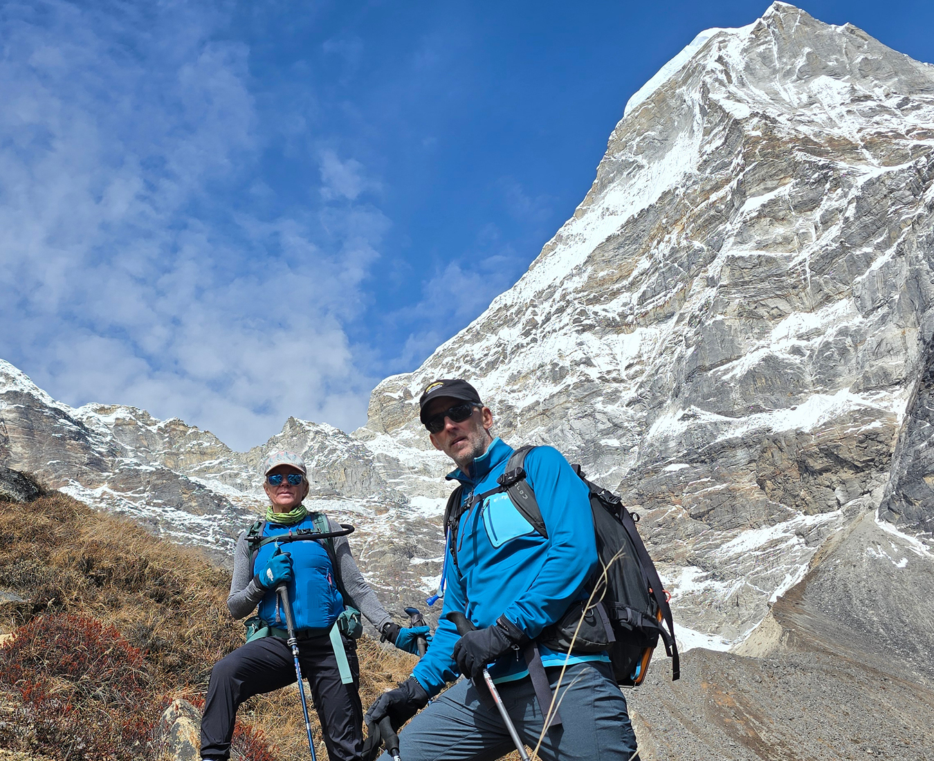 Two climbers wearing blue jackets that match the sky pause on the trail with the giant Kusum Kanguru towering high above them.