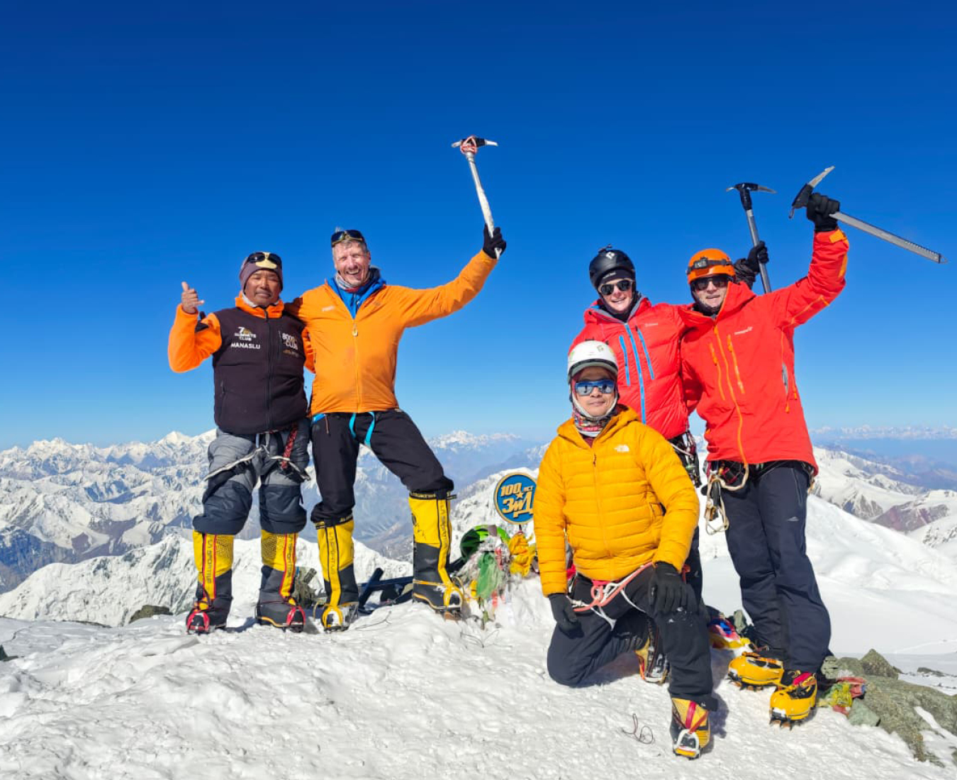 Adventure Consultants Peak Lenin team 2026 stand on the summit of Peak Lenin with their arms and ice axes in the air celebrating their achievement. Brightly coloured yellow, orange and red jackets contrast against a bright blue sky.