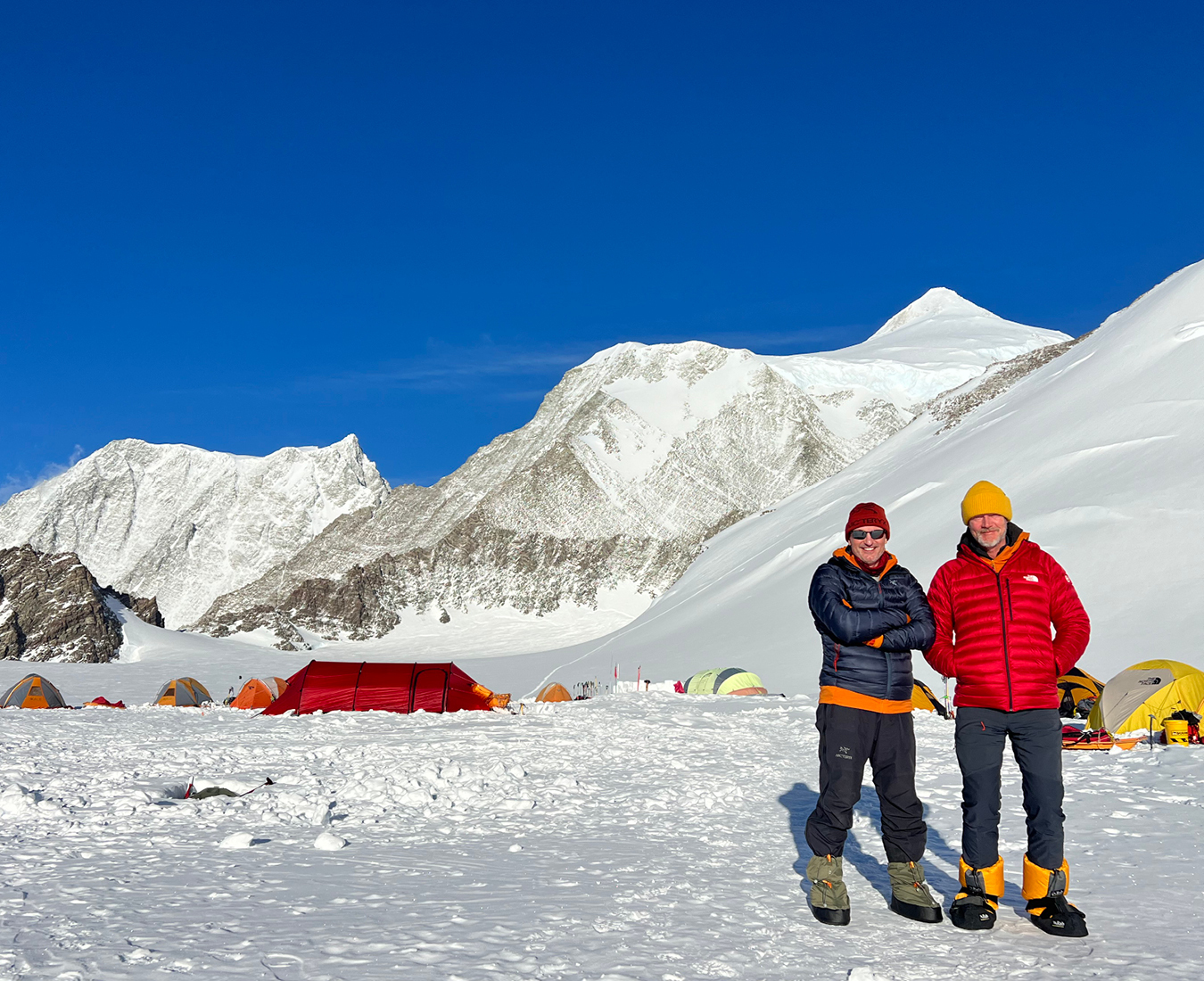 Two climbers relax at Mount Vinson Base Camp ahead of their climb the pointy summit of Mount Shin behind them.