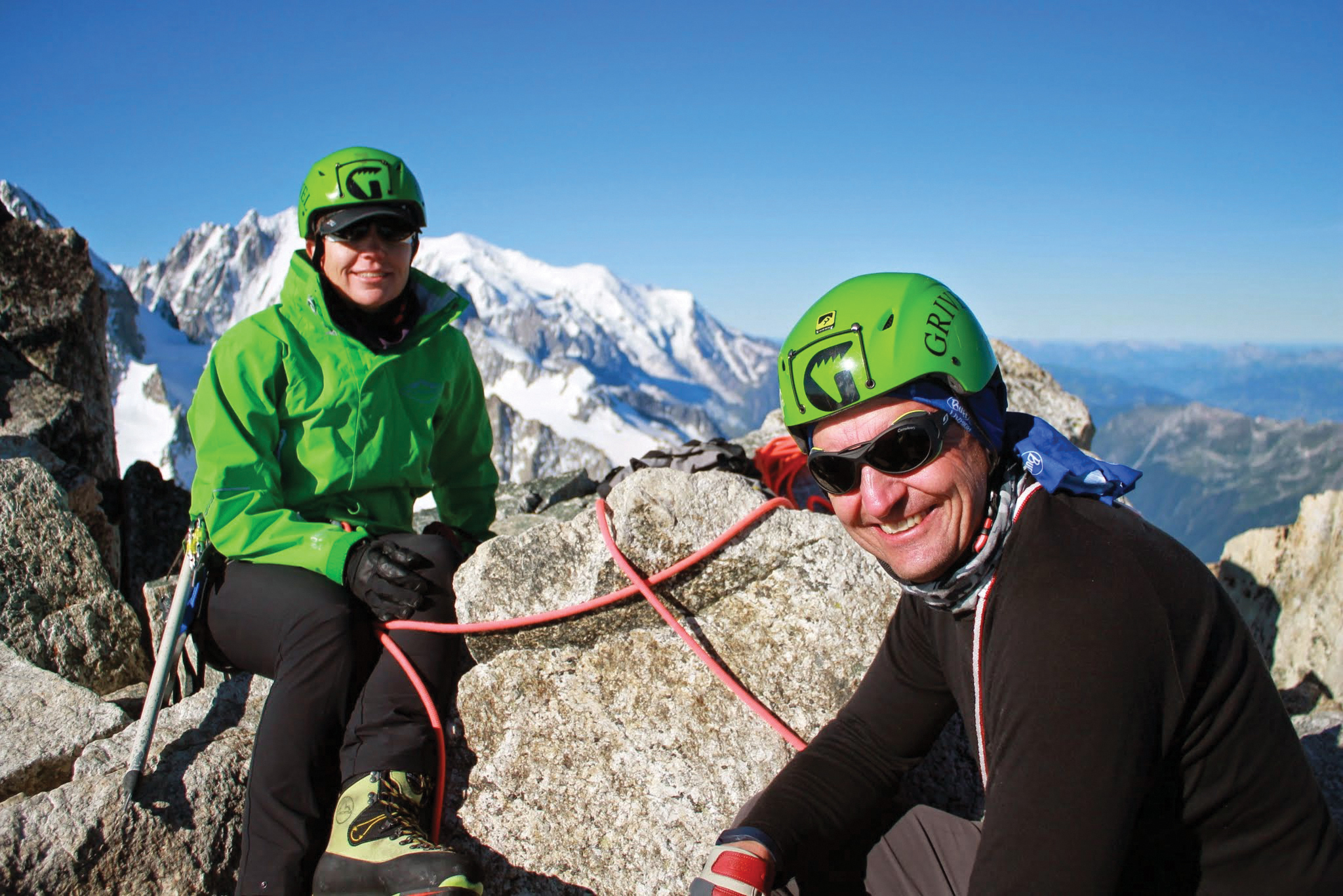 Two climbers in bright green helmets rest on a ridge line high in the European Alps