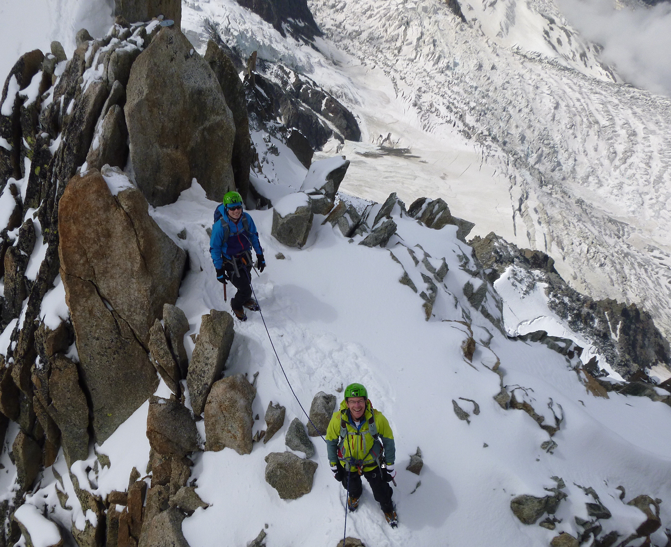 Two climbers roped together high in the alps, steep glaciated terrain surrounds.