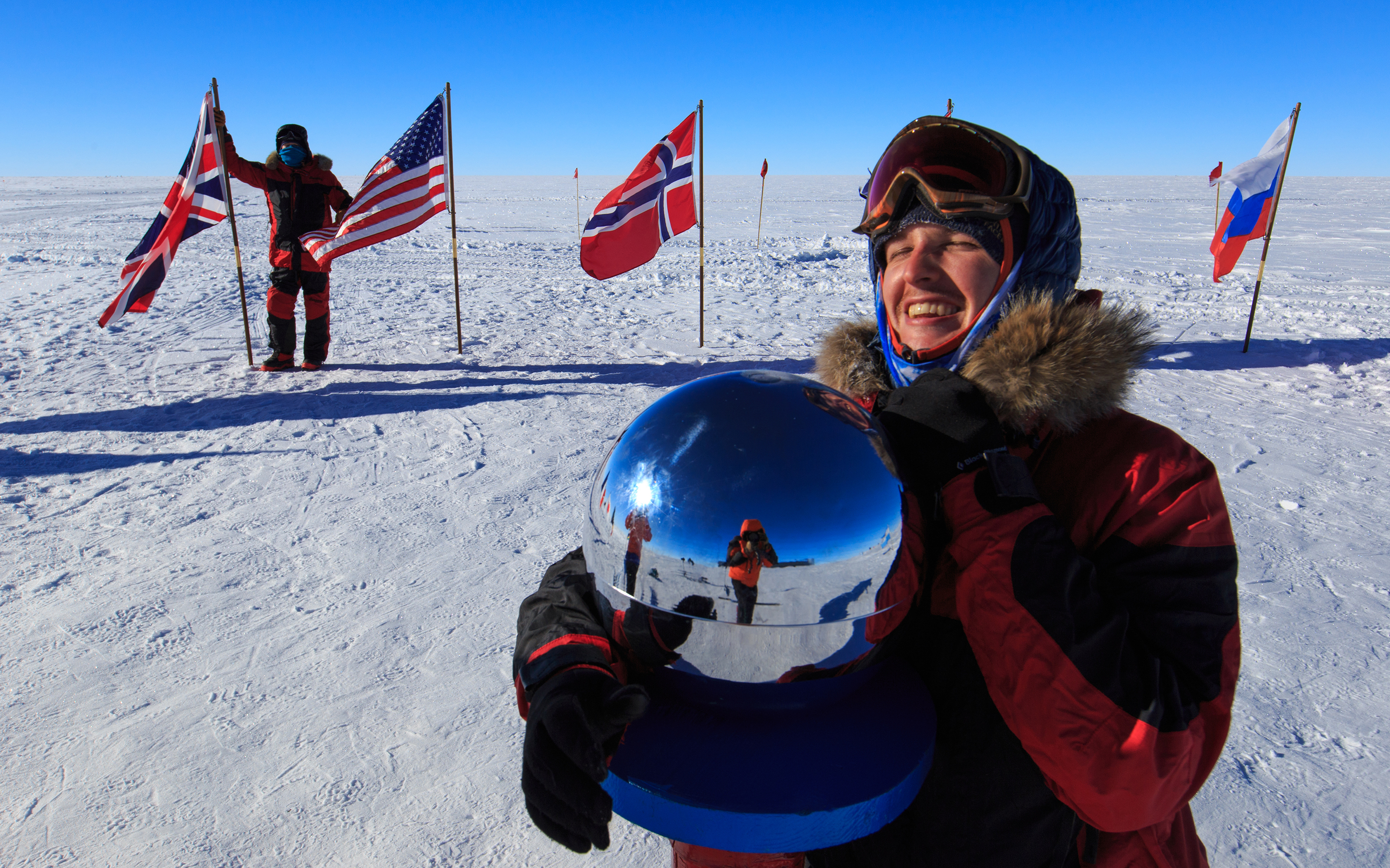 A very jubilant skier hugs the reflective globe at the South Pole while another skier looks on from behind. International flags wave in the breeze.