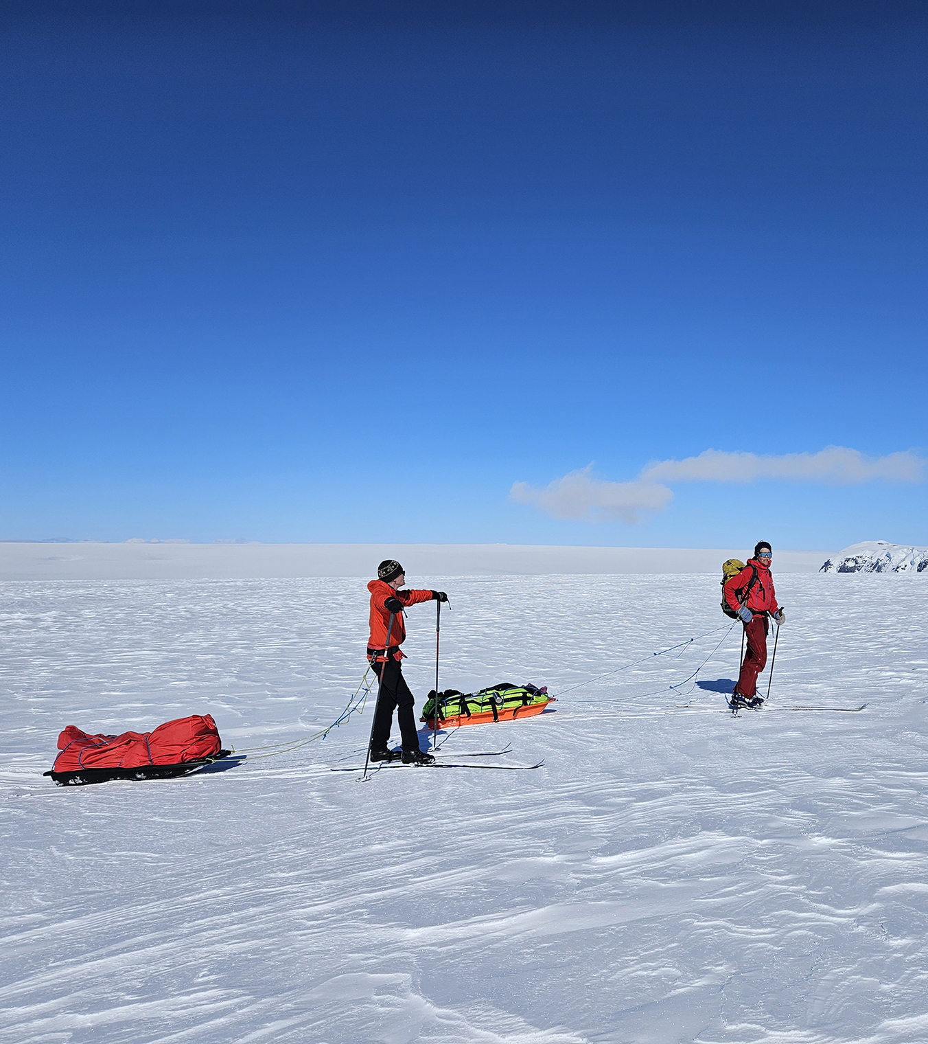 Two skiers hauling sleds pause to take in the enormity of the Greenland icecap, blue skies and ice and snow to the horizon.