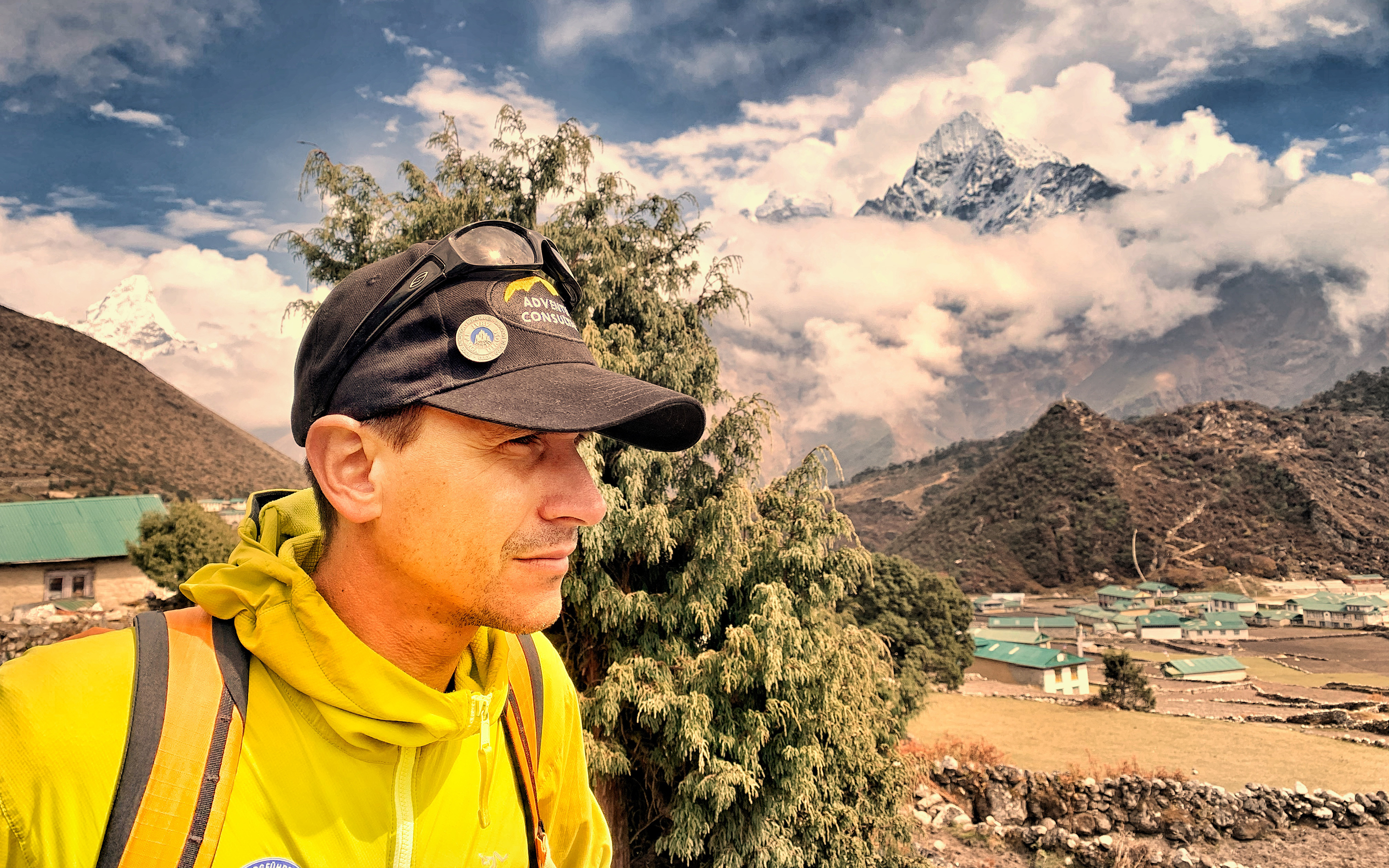 AC IFMGA Mountain & Ski Guide Cosmin Andron stands in front of Mount Ama Dablam, Nepal. Clouds shroud part of the mountain and the village of Khumjung stands directly behind.