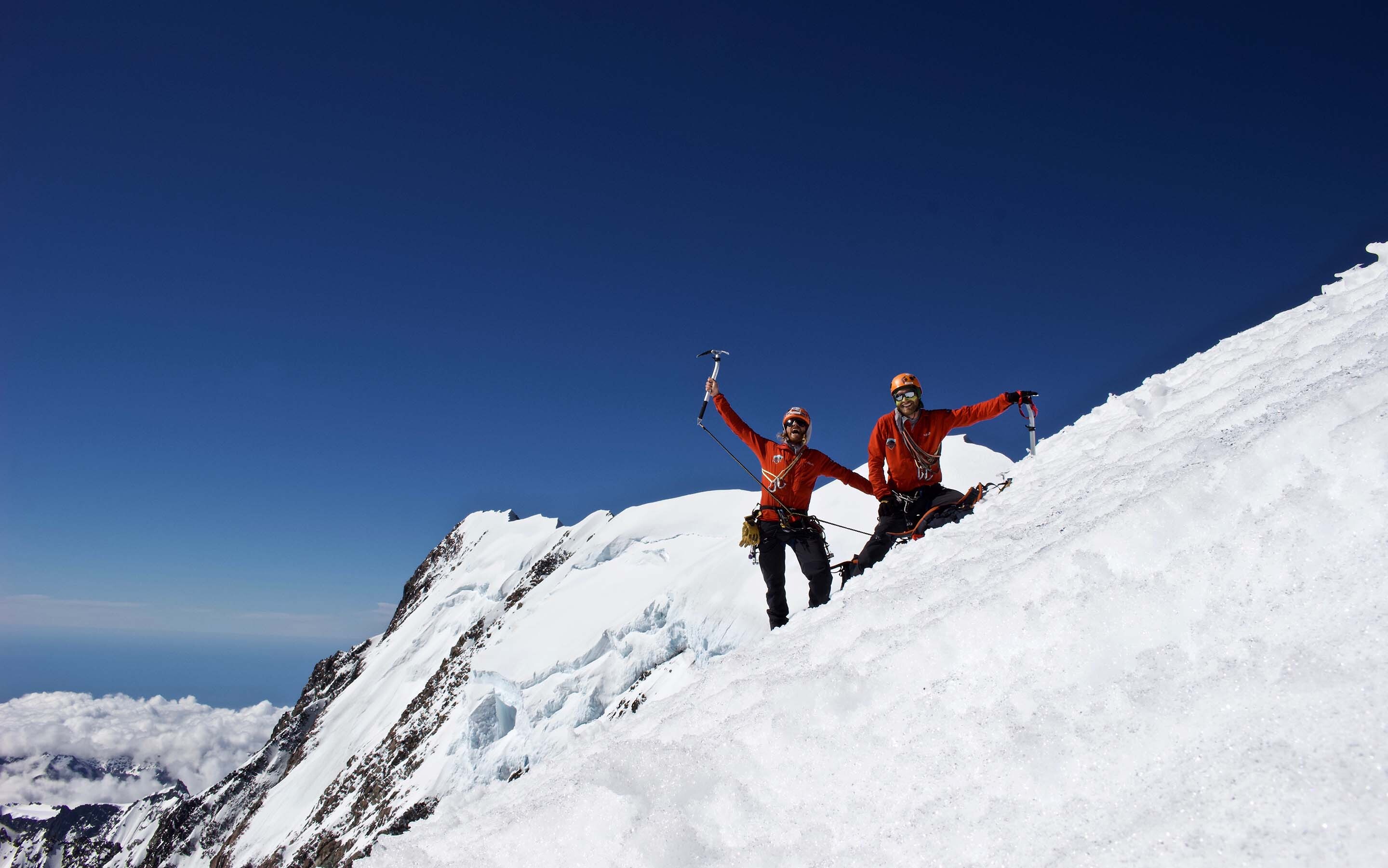 IFMGA Mountain & Ski Guide Dave Lewis celebrates atop a snow covered mountain with bright blue sky contrasting against the white snow.