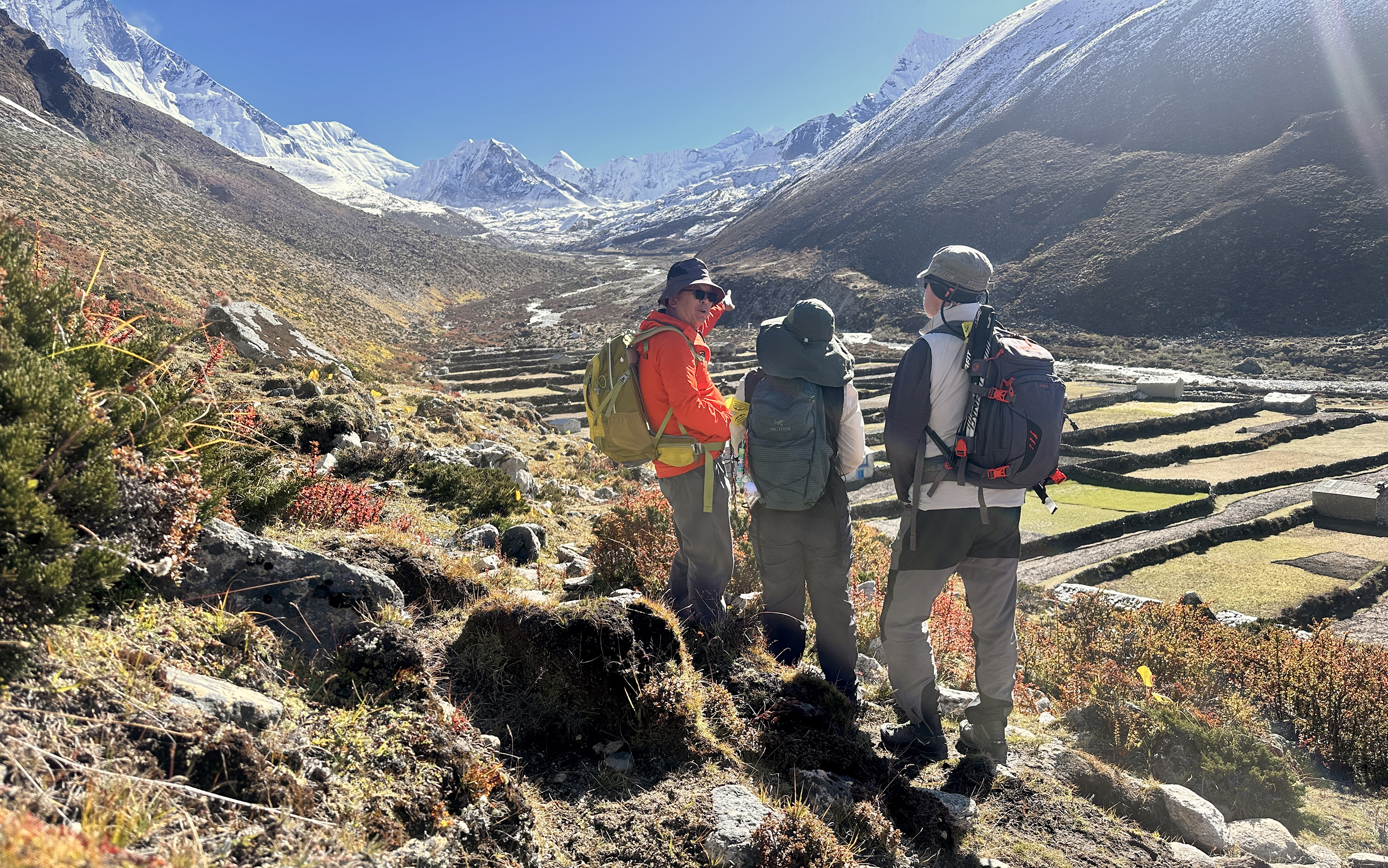 AC Trekking Guide Kami Tenzing Sherpa points out distant peaks (Island Peak, Makalu) to two trekkers, below them the stone-walled fields of DIngboche.