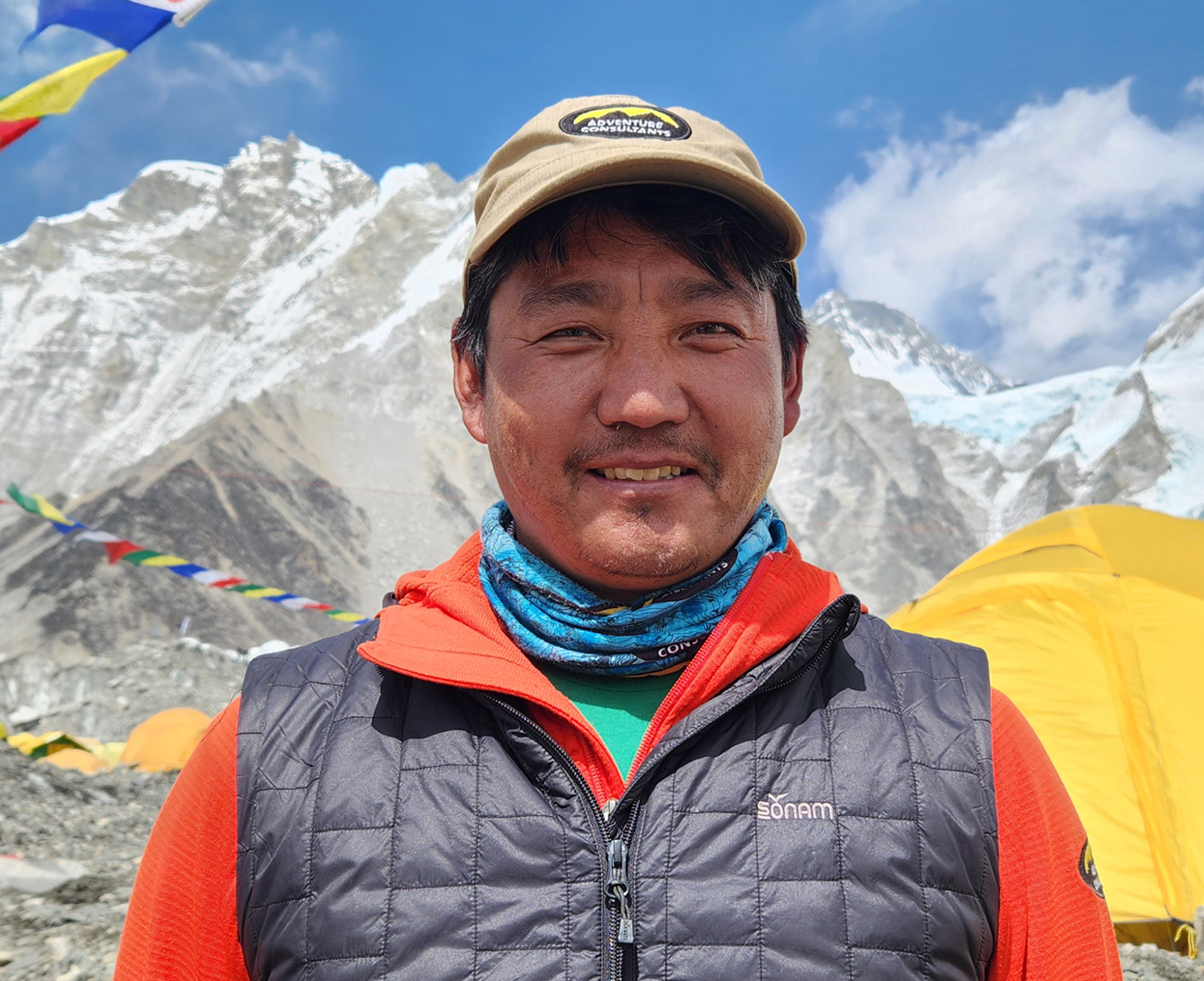 AC Trekking Sherpa Tsering Wangchu at Everest Base Camp, bright yellow tents and prayer flags wave in the breeze behind.