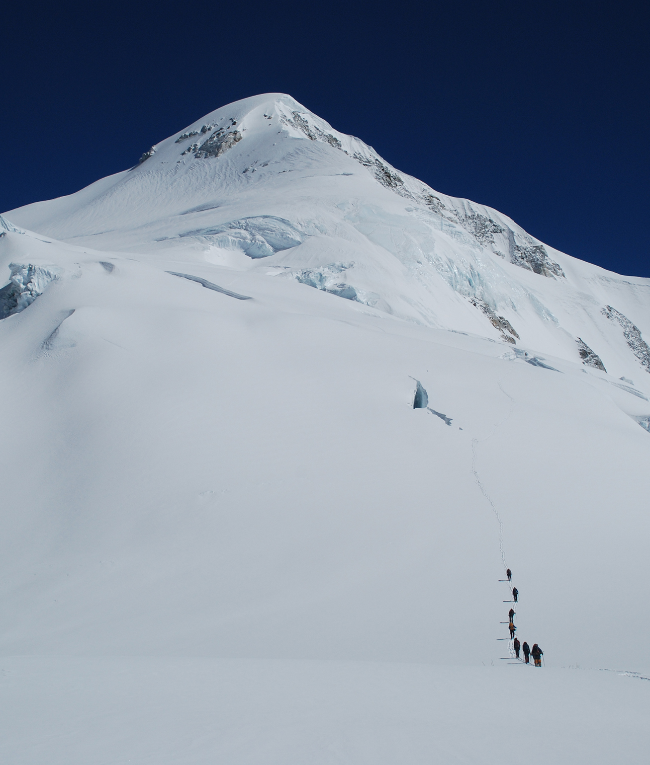 Climbers make their way up a snow covered slope, a large crevasse in front of them and the summit of Baruntse high above