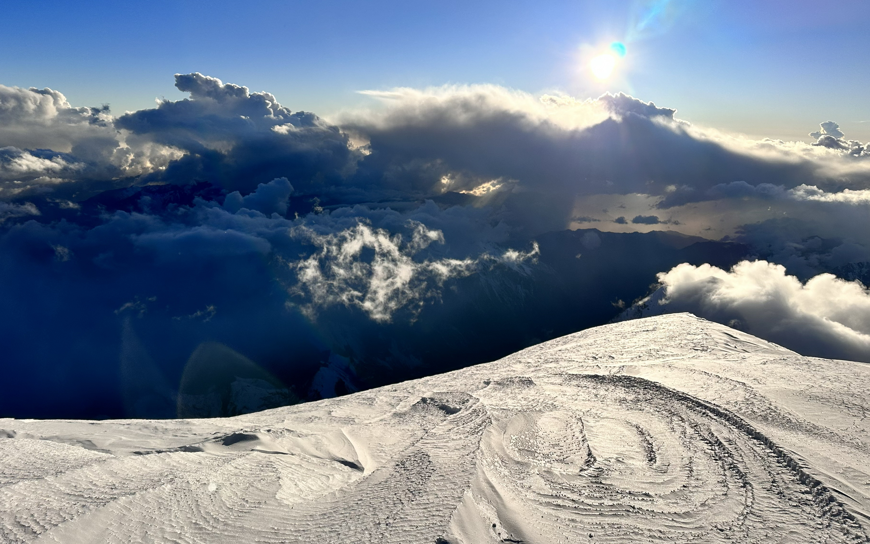 Moody cloud formations in the distance and sun glistening on wind effected snow in the foreground.