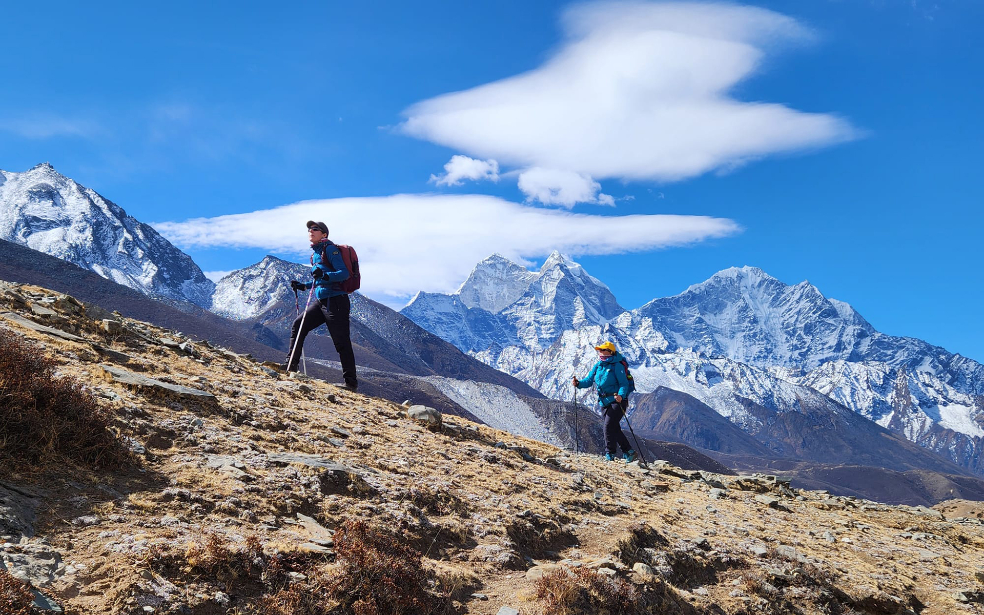 Two trekkers near Pheriche climb a hill with huge mountains in the background.