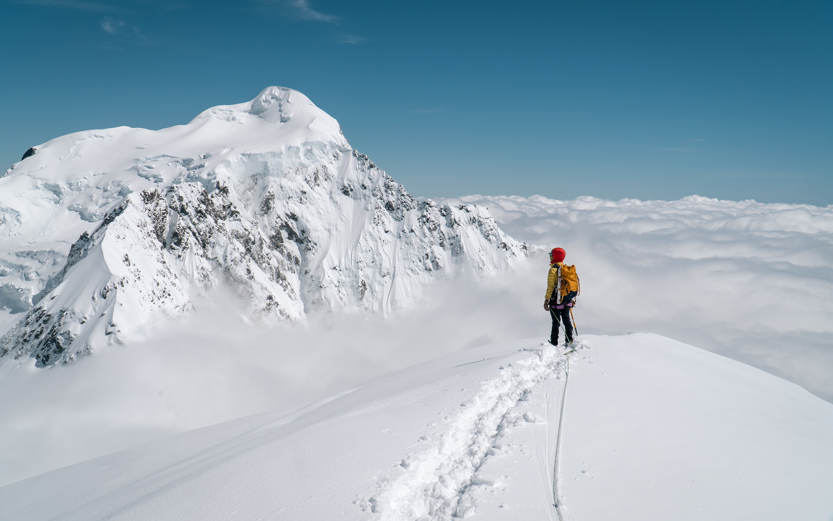 A climber stands atop a snowy peak in New Zealand Southern Alps and looks towards a towering mountain peak, blue sky above and thick cloud below obscuring the view to the mountains below.