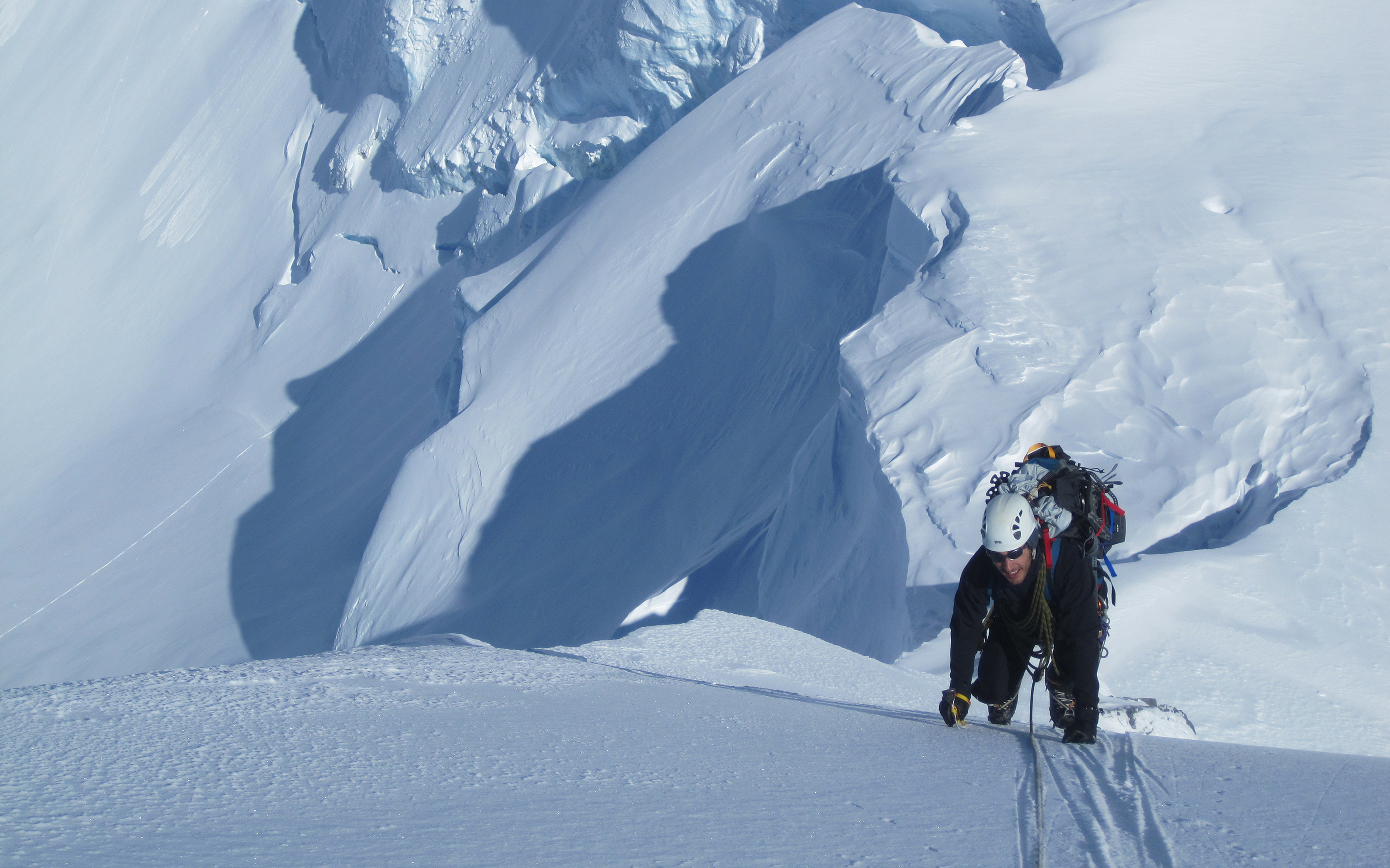 A climber uses ice tools while being belayed up a steep slope in the New Zealand Alps. Large seracs of snow and ice behind.