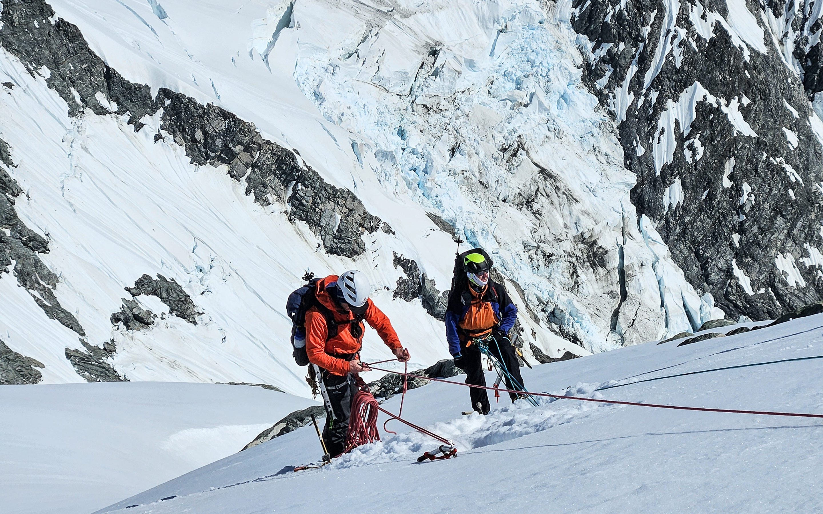 Climbers belay from snow anchors in the New Zealand Alps, crevassed glacial terrain surrounds.