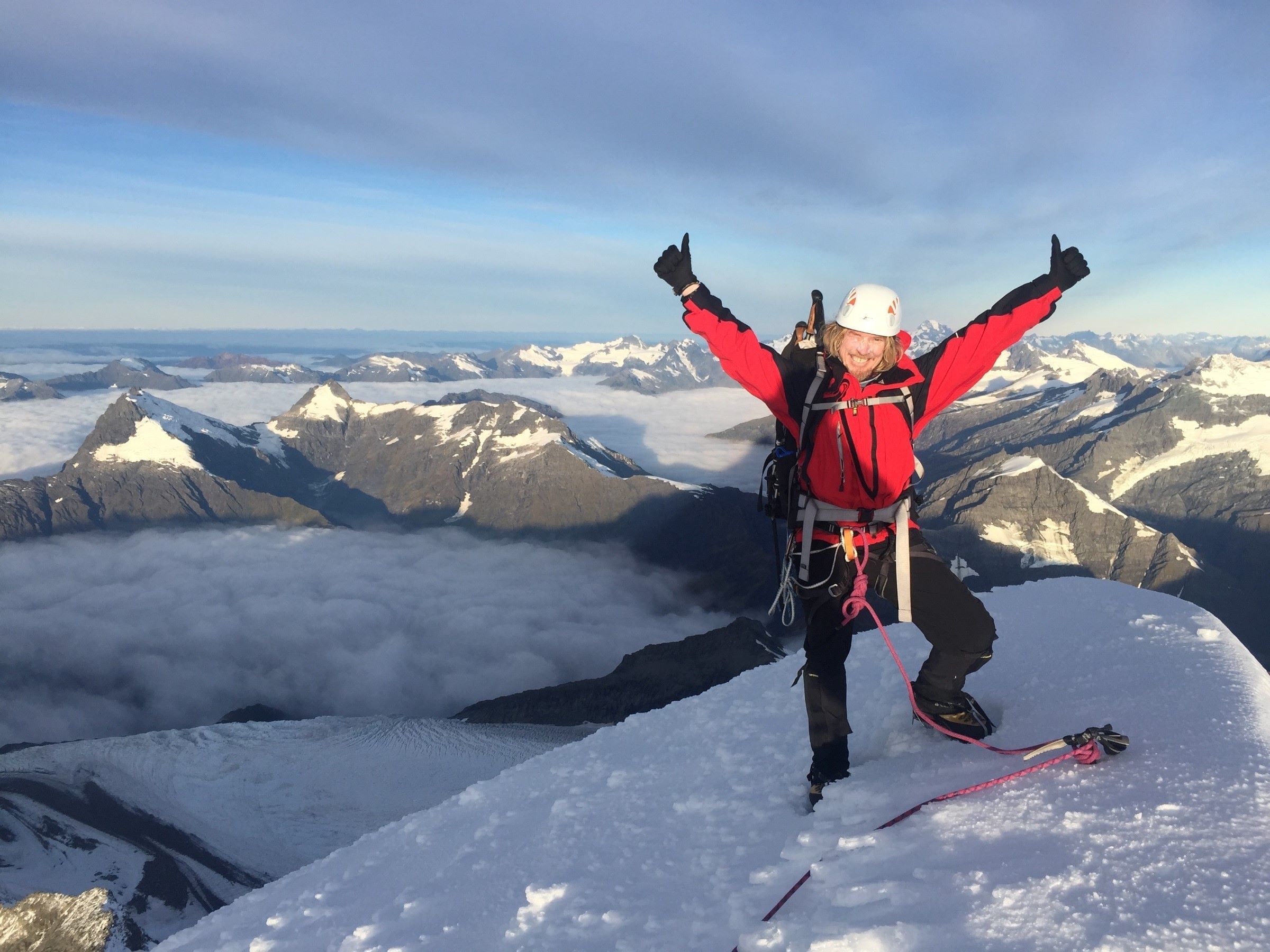 A climber celebrates on the top of a peak in the New Zealand mountains, arms outstretched in elation.