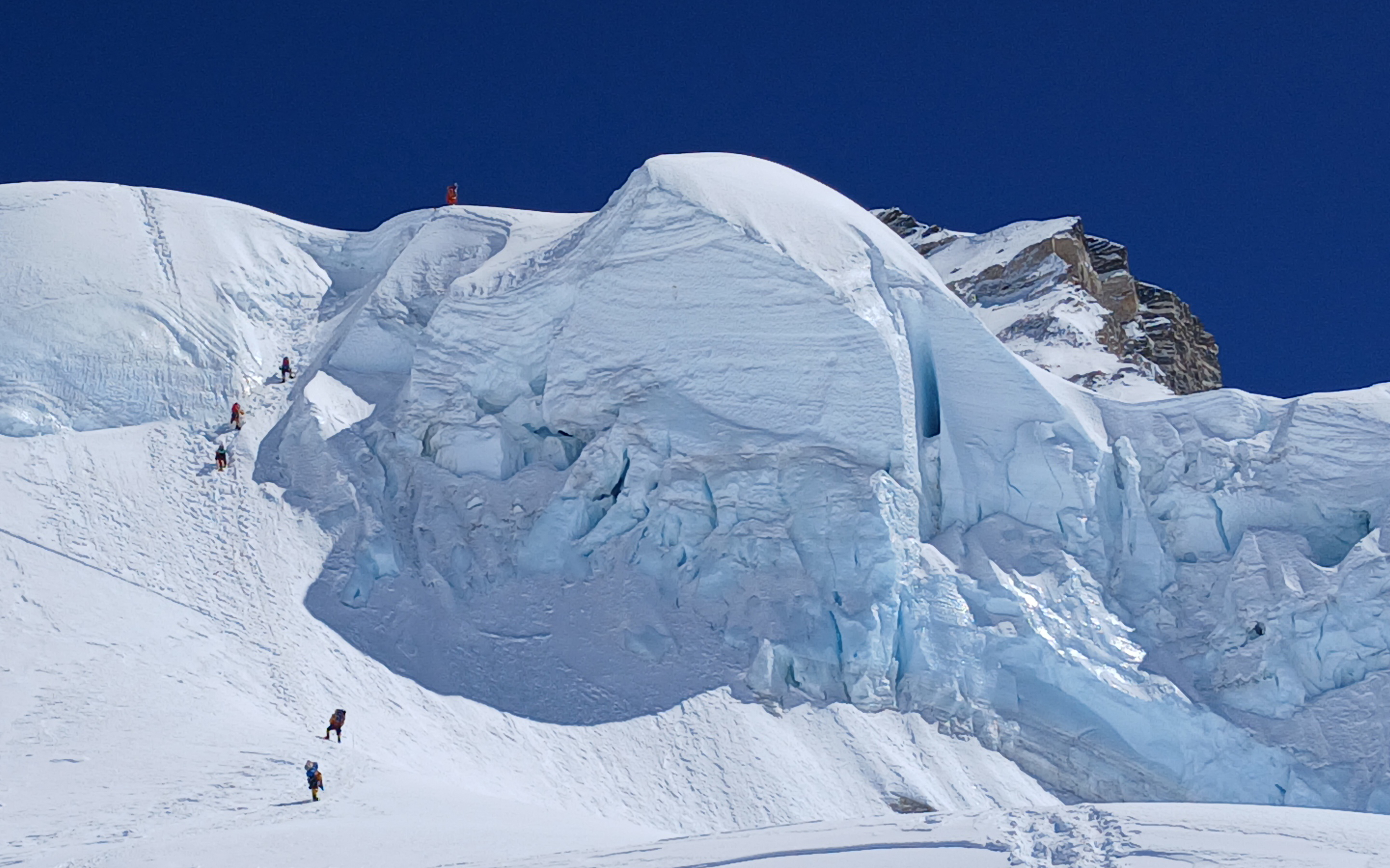 The bulging Cho Oyu ice cliff, the crux of the climb dwarfs climbers as they make their way upwards. Bright blue sky above.