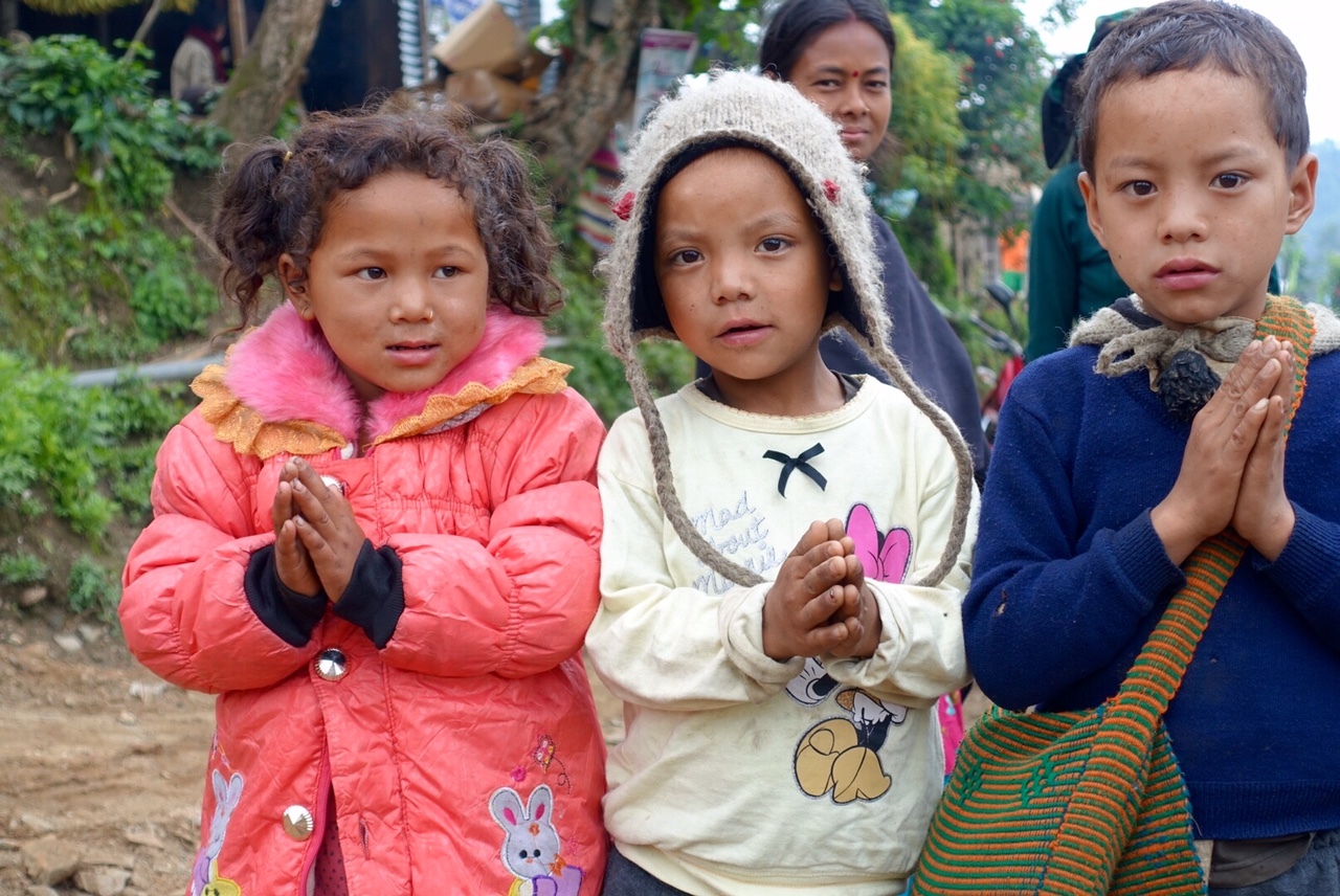 Three young children stand on a village path with hands pressed together in greeting, watching visitors quietly as adults gather behind them