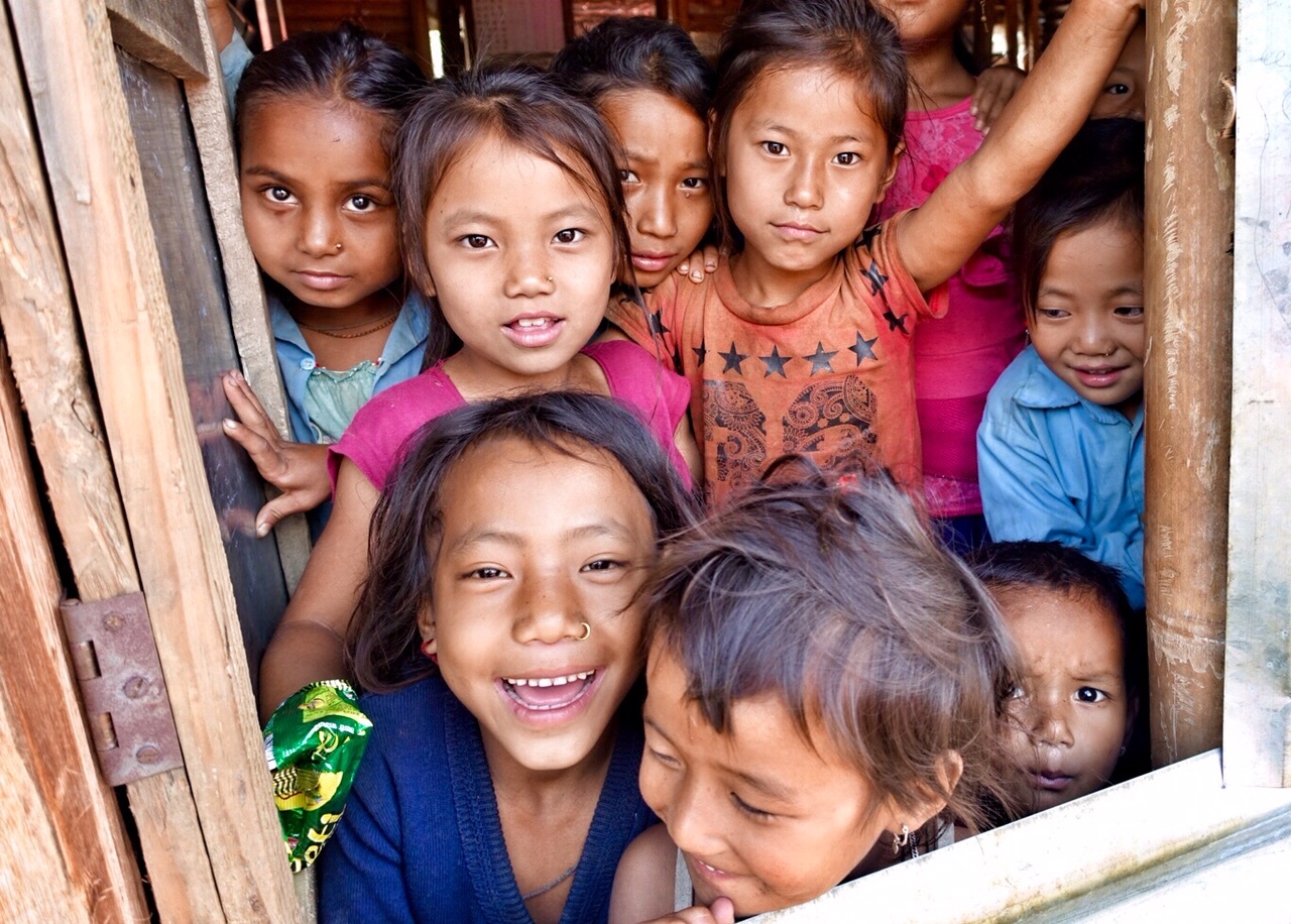 A group of smiling children crowd a wooden doorway, peering out together, their faces close, curious and full of warmth despite simple surroundings.
