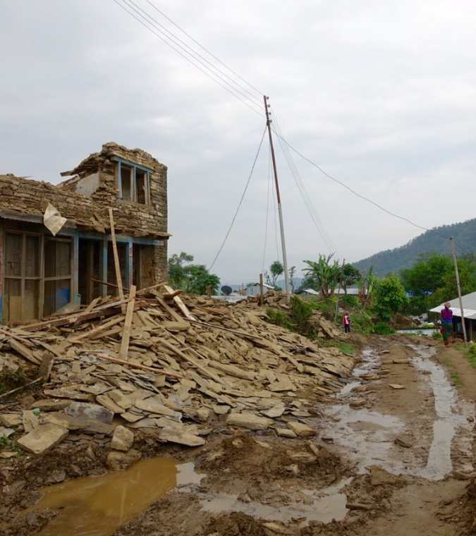 A damaged village in Gorkha, Nepal, where collapsed stone houses line a muddy road, showing the lasting impact of the 2015 earthquake.