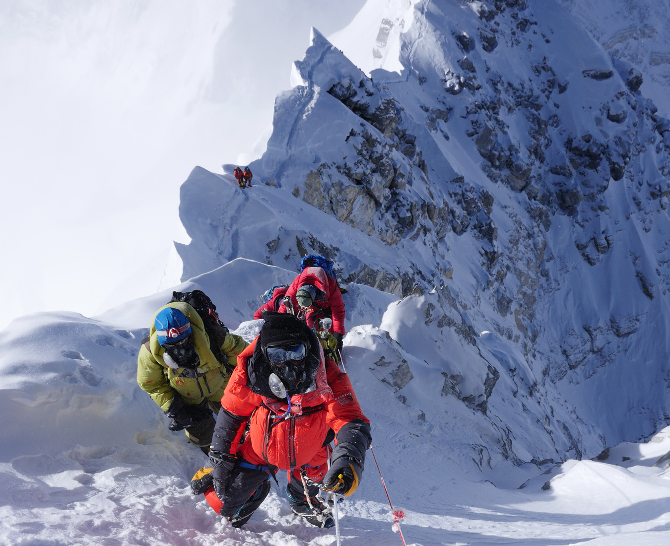 Climbers high on the steep summit ridge of Mount Everest, oxygen masks obscure their faces as they climb fixed ropes.