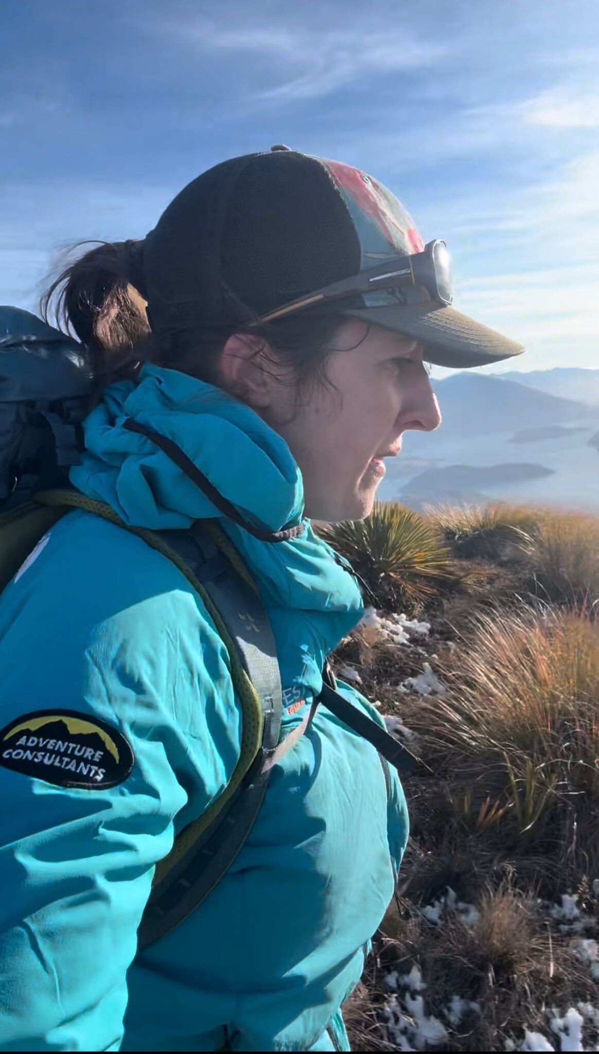 Adventure Consultants General Manager Caroline Ogle training on a mountain ridge in New Zealand, wearing an AC-branded teal jacket, cap, and glacier glasses. Layers of cloud-covered ranges stretch out behind her.