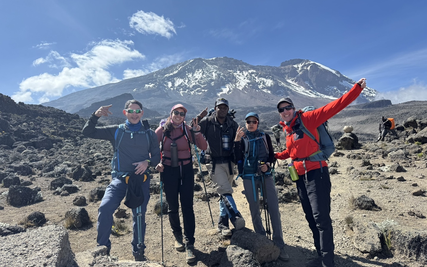 A group of five trekkers pose on the rocky moorland of Kilimanjaro's Shira-Barafu route, with the snow-capped summit visible behind them. Everyone is grinning and throwing up peace signs and pointed fingers. A local guide stands at centre, backpack loaded.