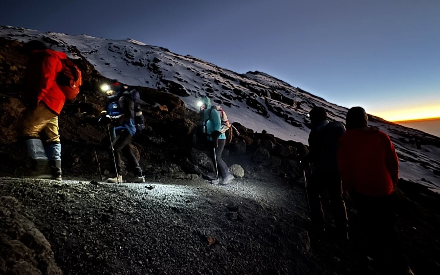 Trekkers wearing headlamps climb a steep scree slope in darkness on Kilimanjaro's summit night, trekking poles in hand. The horizon glows amber and gold as the first light of sunrise appears over the clouds far below.
