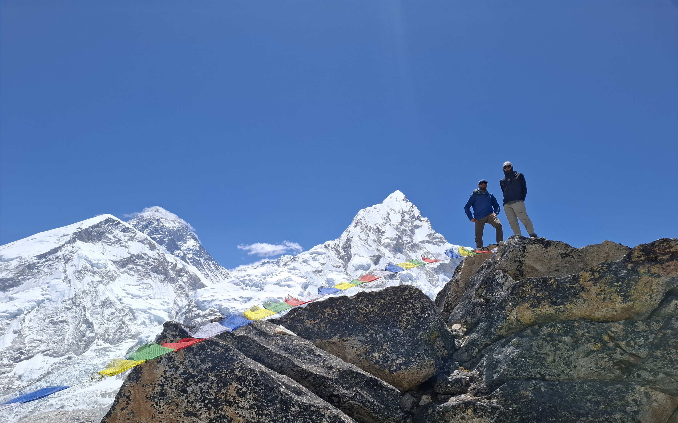 Two climbers stand at Pumori Camp 1 with Everest, Lhotse and Nuptse in view behind them, bright blue sky above.