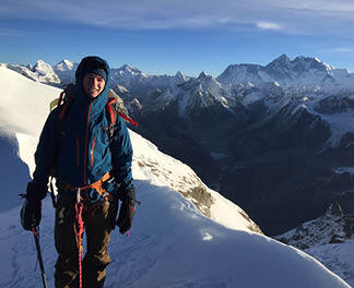 Spectacular views from the summit of Mera Peak, a climber stands with distant 8,000m peaks behind in clear blue sky weather.