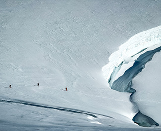 Three climbers traverse across a glacier towards a snow bridge that bridges the gap of an open and deep crevasse.