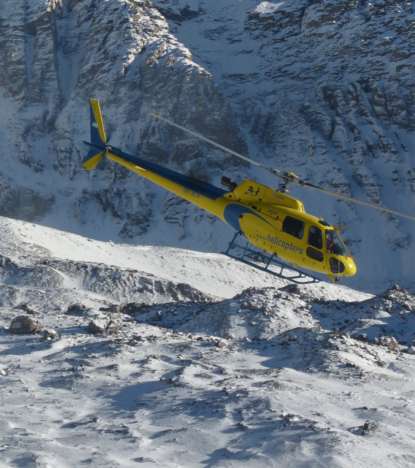A yellow helicopter takes off from Aconcagua Base Camp, snow on the ground and steep sided mountains surround.