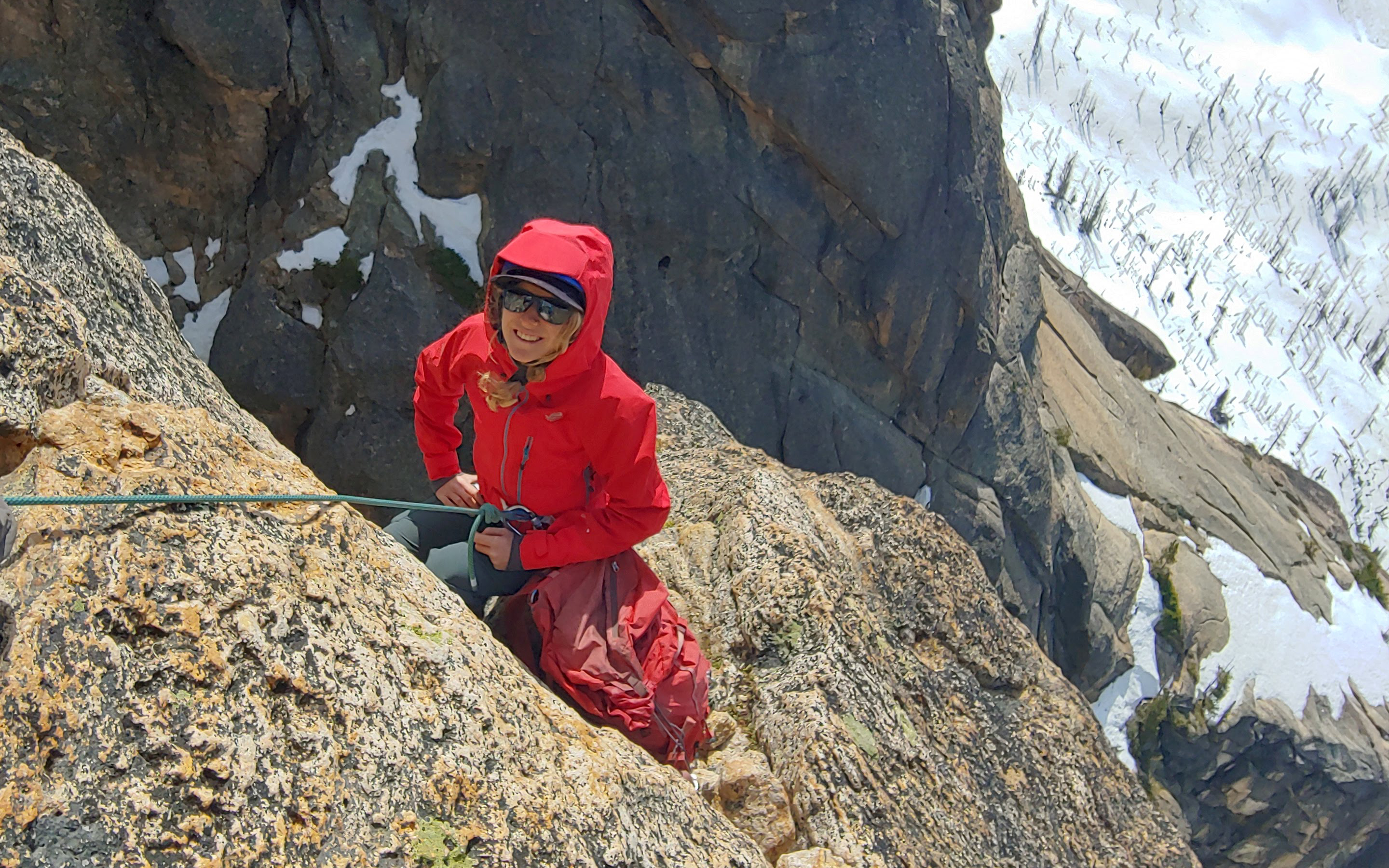 A climber stands out against the landscape wearing a bright red jacket, with snow below they look up towards their guide above.