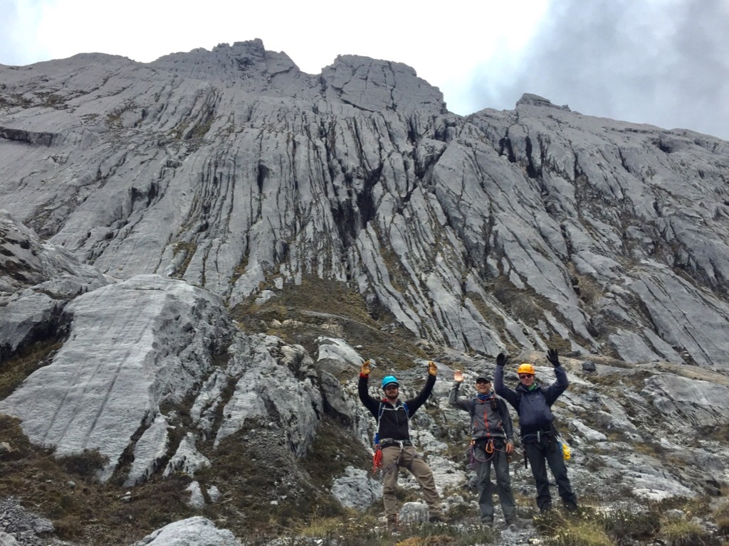 An Adventure Consultants team stands at the base of Carstensz Pyramid, the imposing limestone escarpment towering above.