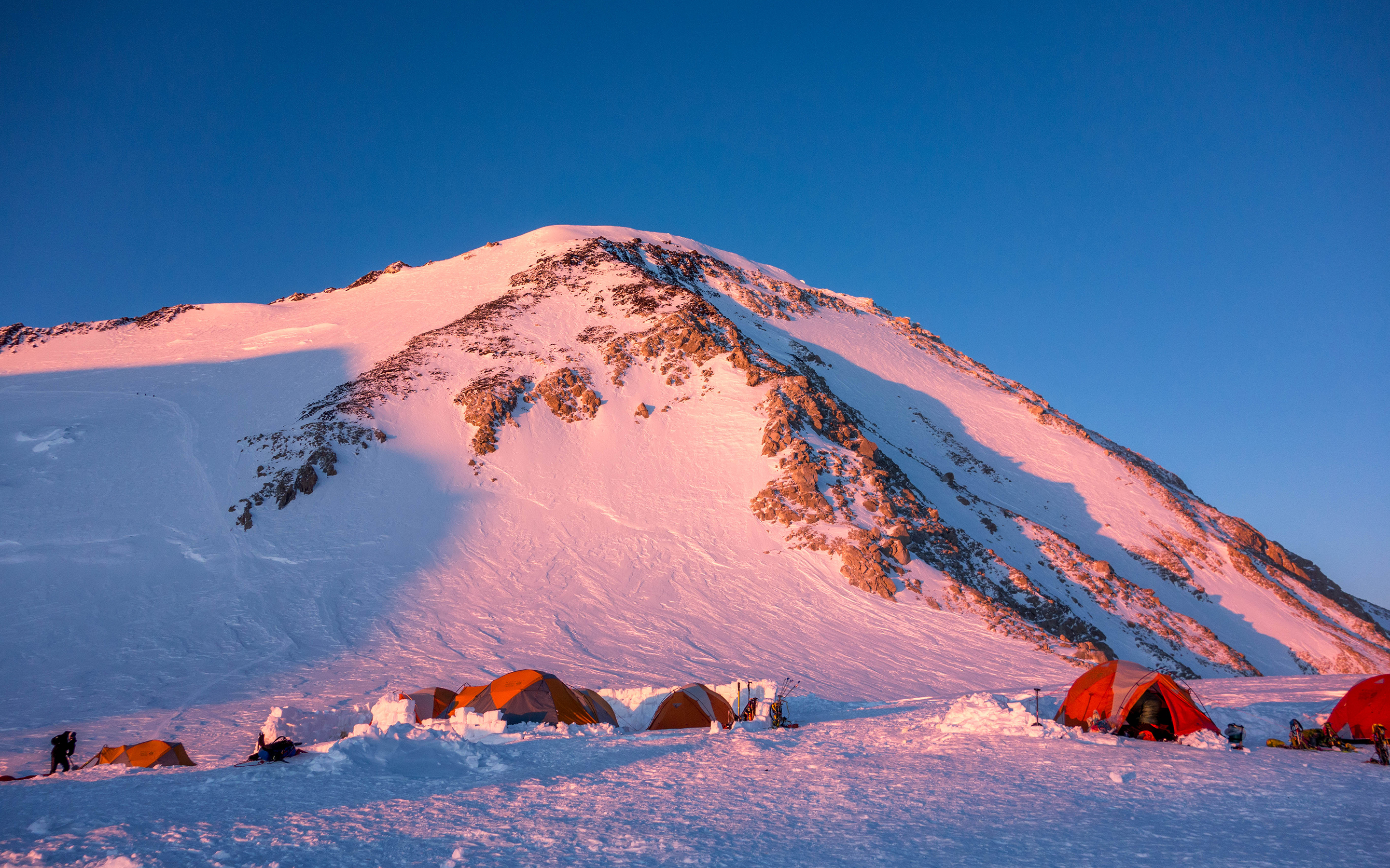 Evening light glows pink on the slopes of Denali with tents surrounded by snow walls in the foreground