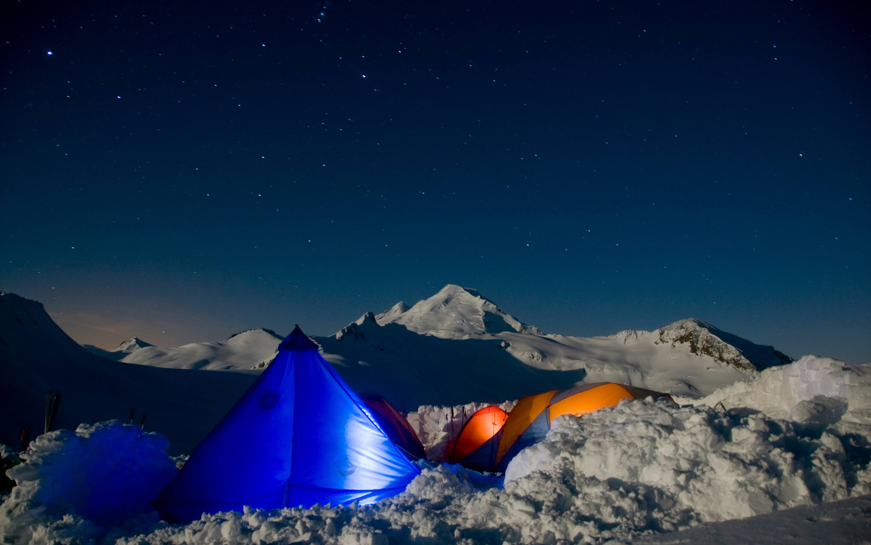 Tents pitched in snow illuminated by torch light against the night sky.