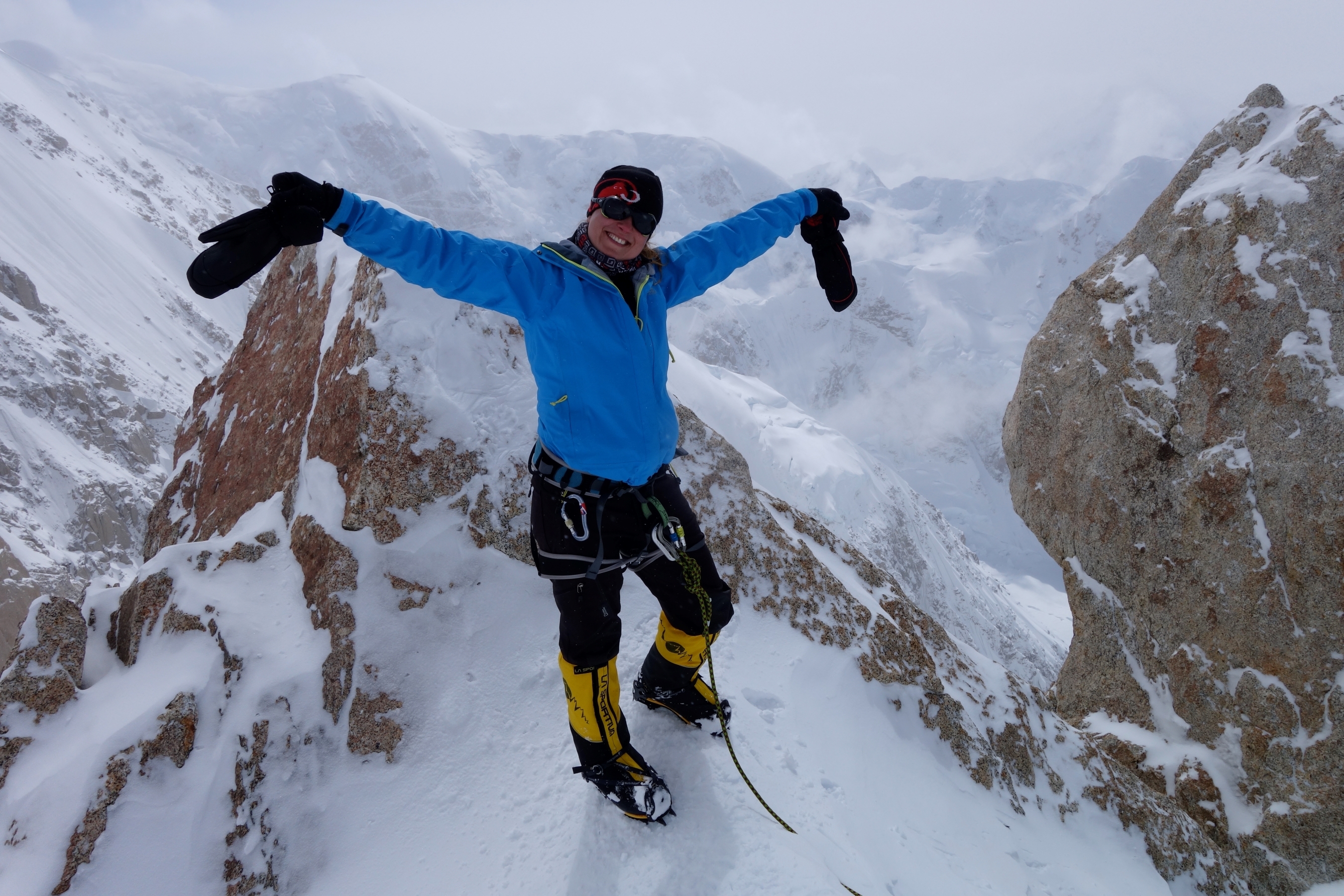 A climber celebrates on the summit of Denali, peaks and cloud behind.